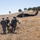 Military personnel in camouflage uniforms are carrying a casualty on a stretcher across a dry, grassy field toward a waiting medical evacuation helicopter. The helicopter, marked with a red cross indicating medical/MEDEVAC designation, is landed on a hillside in the background. This appears to be either a combat casualty evacuation operation or a military training exercise.