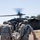 Military personnel in U.S. Army combat uniforms are carrying a casualty on a litter (stretcher) toward a UH-60 Black Hawk helicopter with rotors spinning. The scene appears to be a medical evacuation (MEDEVAC) operation or training exercise conducted in an open, dry grassland field. Several soldiers are involved in loading the casualty while crew members are visible near the helicopter's open side door.
