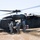 A military medical evacuation (MEDEVAC) operation is underway in an open field with dry grass. Several soldiers in ACU (Army Combat Uniform) camouflage are loading a casualty on a litter into a UH-60 Black Hawk helicopter marked with a Red Cross symbol, indicating its medical role. The helicopter's rotors are spinning, suggesting an active or imminent departure.