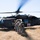 A military medical evacuation (MEDEVAC) helicopter, identified by the prominent Red Cross symbol, is landed in a dry grassy field. Several soldiers in camouflage uniforms and helmets are disembarking or standing near the aircraft. The scene appears to be either a training exercise or an active MEDEVAC operation in a rural, arid environment.