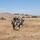 The image shows military personnel in a field training exercise, with two soldiers carrying a third soldier who appears to be simulating a casualty. The scene takes place in an arid, open grassland terrain with rolling hills in the background. A fourth soldier stands nearby observing, and a military vehicle (likely a HMMWV/Humvee) is visible in the background.