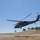 A military medical helicopter, identifiable by the red cross marking on its fuselage, is flying at low altitude over dry, golden grassland terrain. The helicopter appears to be a UH-60 Black Hawk configured for medevac operations. The background shows scattered oak trees on rolling hills under a clear blue sky, suggesting a warm, dry environment consistent with California or similar terrain.