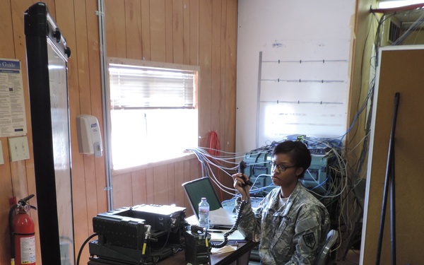 A U.S. Army soldier in ACU (Army Combat Uniform) is seated at a communications workstation inside a temporary or field operations room. The soldier is holding a radio handset and appears to be operating military communications equipment. The room contains multiple electronic devices, cables, a laptop, and various field communication gear.