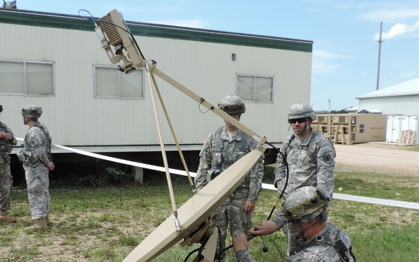 Several U.S. Army soldiers in ACU (Army Combat Uniform) camouflage are setting up a portable satellite communications dish on a grassy area near military-style modular buildings. The equipment appears to be a man-portable satellite terminal mounted on a tan/beige base platform with the identifier '4320' visible. The scene suggests a field training exercise or operational communications setup.