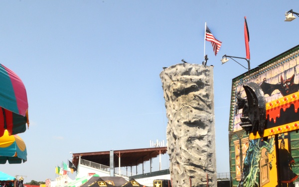 The image shows an outdoor fairground or carnival on a clear, sunny day. Visible attractions include a rock climbing wall, a haunted house-style attraction with horror-themed artwork, colorful vendor umbrellas, and a U.S. Army recruitment booth. Several fairgoers are visible walking along a paved midway.