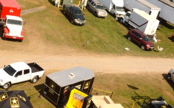 An aerial view of a U.S. Army recruitment and exhibition booth at what appears to be an outdoor fair or event. The display includes branded tents, a military HMMWV (Humvee), interactive stations, and a rock climbing wall. The setup prominently features GoArmy.com branding and recruitment contact information.