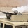 A soldier in full combat gear is operating a crew-served machine gun mounted on the turret of a tan/desert-colored HMMWV (Humvee). The weapon appears to have just been fired, as evidenced by a large white smoke cloud visible in the background. The setting appears to be an arid, desert environment consistent with a training range or operational area.