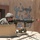 A U.S. Army soldier in ACU (Army Combat Uniform) is positioned at a vehicle-mounted M240 machine gun, appearing to be on overwatch or patrol duty. The soldier is wearing a combat helmet and ballistic eyewear, positioned behind a vehicle gun shield. Arabic/Dari script graffiti is visible on a tan mud-brick wall in the background, suggesting a Middle Eastern or Central Asian operational environment.