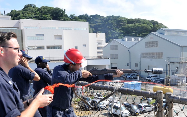 Three naval personnel in dark blue uniforms are positioned on the deck of a military vessel, with one individual wearing a red safety helmet and aiming a line-throwing gun toward the pier. The activity appears to be a mooring or line-handling drill conducted at a naval facility, likely in Japan based on the visible vehicles and architectural style. White industrial and administrative buildings, a parking lot with numerous vehicles, and tree-covered hills are visible in the background.
