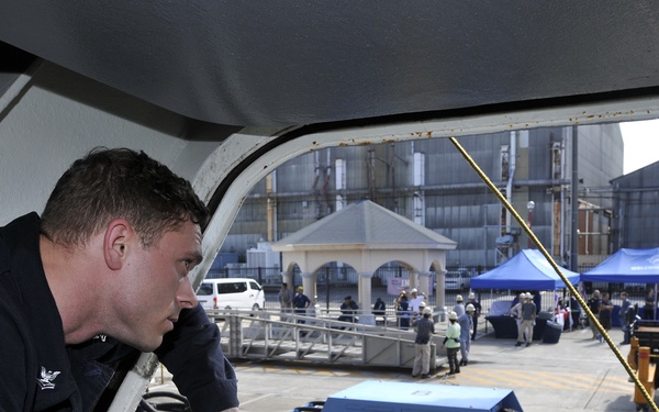 A male individual wearing a dark naval/military uniform is leaning out of an elevated opening or hatch, looking down at a pier or dock area below. The scene appears to be at a naval facility or port, with equipment, cables, and a gathering of people visible on the dock. Blue tents and a small gazebo structure are visible in the background, suggesting an official event or ceremony.