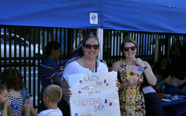 A military homecoming event where a woman holds a handmade sign reading 'Outta my way, I get my HUSBAND BACK today... Welcome Home Chief GROW.' The gathering takes place under a blue canopy tent with children and adults present, and another woman holds a small American flag. The atmosphere is celebratory and patriotic.
