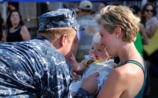 A U.S. Navy sailor in digital camouflage uniform is reuniting with a woman holding an infant, likely at a homecoming event. The sailor is leaning in to interact with the baby, while the woman smiles warmly. A crowd of onlookers is visible in the background, consistent with a military homecoming ceremony.