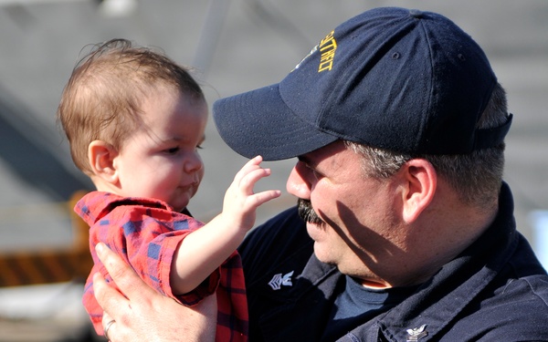 A military servicemember in dark navy uniform and cap is holding a baby dressed in a red plaid outfit, while another child in a black-and-white striped shirt stands in front of him. The scene appears to be a homecoming or reunion event, likely at a naval base or pier. The emotional interaction between the servicemember and the infant suggests a family reunion after a deployment.