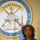 A woman in a teal blazer stands at a wooden podium, appearing to speak or present. Behind her is a large official seal reading 'NAVSCIATTS' (Naval Small Craft Instruction and Technical Training School), along with a blue flag partially visible. She wears a necklace and a small lapel pin.