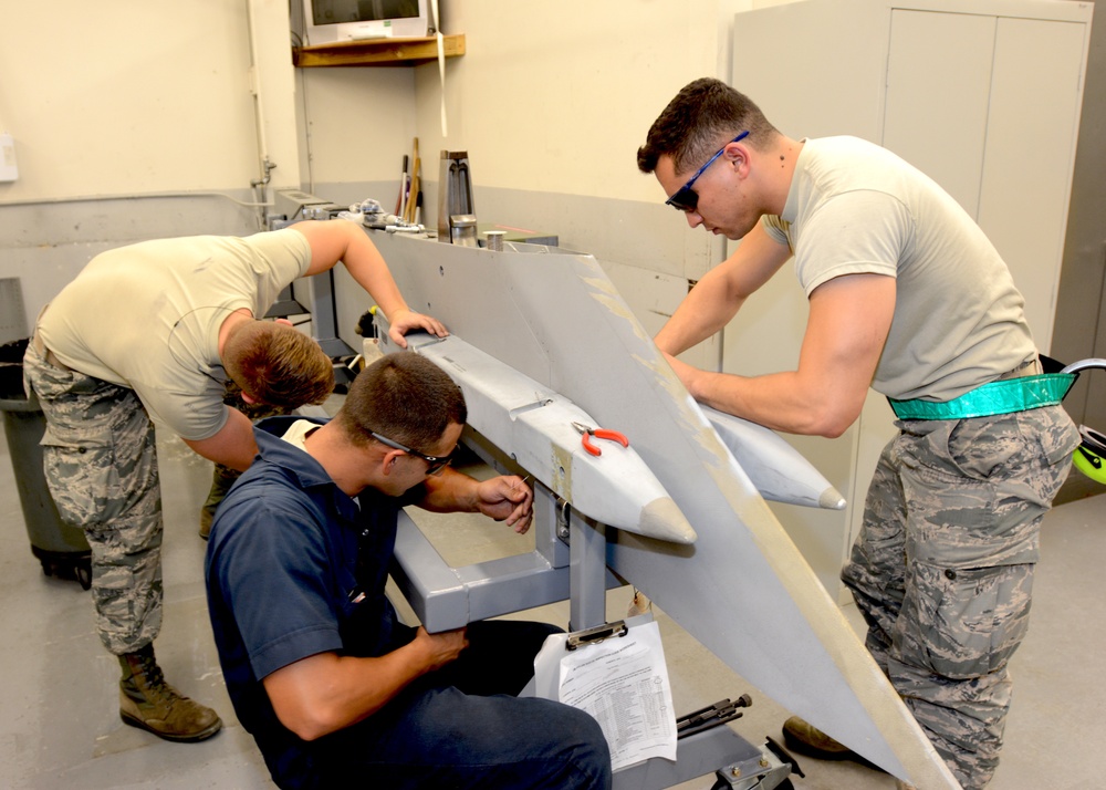 Three military personnel, appearing to be U.S. Air Force members based on their ABU (Airman Battle Uniform) camouflage pattern, are working on what appears to be an air-to-air or air-to-ground missile or munition component mounted on a maintenance stand. One individual in civilian clothing is crouching and working on the lower section while referencing a document checklist, while two uniformed airmen assist from above. The setting is an indoor maintenance or weapons shop facility.