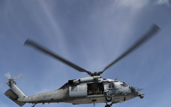 A U.S. Navy helicopter, identifiable by 'NAVY' markings on the fuselage, hovers over a ship's flight deck while conducting a vertical replenishment (VERTREP) operation. A cargo net loaded with supplies is suspended beneath the helicopter via a hoist cable. A second vessel is visible in the background, likely the supply ship from which cargo is being transferred.