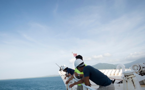 Two crew members wearing hard hats and safety vests are leaning over the railing of a large vessel, looking down toward the waterline. The ship appears to be a cargo or military vessel navigating calm blue ocean waters near a mountainous coastline. An American flag is visible flying from a mast in the background.