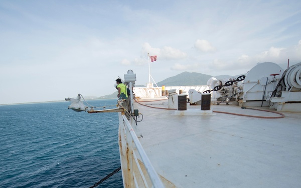 The image shows the bow deck of a large white vessel at sea, with mountainous coastline visible in the background. Two crew members are visible on deck, one wearing a high-visibility yellow vest near the port bow and another near winch equipment on the starboard side. An American flag is flying from a mast, and anchor chain equipment is prominently visible along the ship's rail.