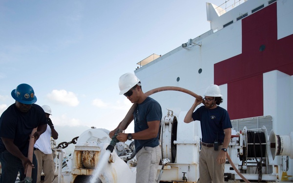 Three workers in hard hats are performing deck maintenance aboard a large white hospital ship, identifiable by the prominent red cross markings on its superstructure. The crew members are using high-pressure water hoses to clean the ship's forward deck area, which shows signs of rust and grime. The scene appears to be in a tropical or subtropical location based on the vegetation visible in the background.
