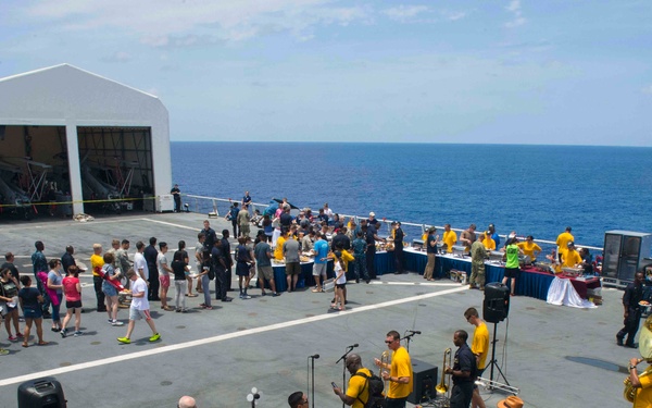 A live band wearing matching yellow shirts performs on the flight deck of a naval vessel during what appears to be a morale or recreational event. A large crowd of people, including civilians and military personnel, are lined up at food service tables in the background. The event takes place at sea, with open ocean visible on the horizon.