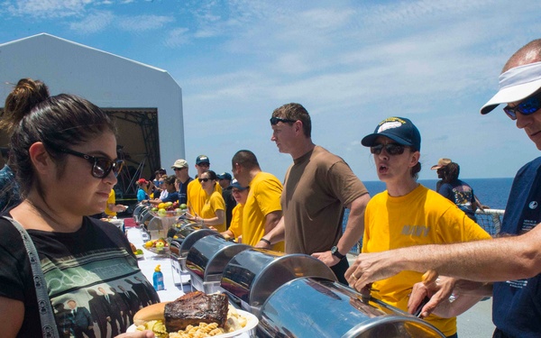 An outdoor buffet event is taking place on the deck of a naval vessel at sea. Personnel in yellow t-shirts are serving food from chafing dishes along a white-tablecloth buffet line to guests. The ocean is visible in the background under a partly cloudy sky.