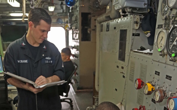 Two U.S. Navy personnel are working inside what appears to be a ship's engineering or mechanical control room. One sailor, identified by name tape as 'MCRANIE,' stands holding a clipboard and observing, while a second sailor crouches down adjusting valves on a large control panel. A third person is visible seated in the background at a workstation.