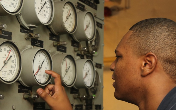 A man in a dark blue uniform is examining and pointing at pressure gauges on an industrial control panel. The panel contains multiple analog gauges, digital readouts, and labeled instrument clusters, suggesting a military or industrial engineering environment. The individual appears to be conducting an inspection or monitoring system readings.