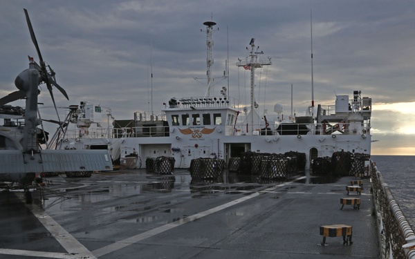 The image shows the wet flight deck of a naval vessel at sea, with a helicopter parked and secured on the port side. Alongside the ship is a second vessel, appearing to be a replenishment or auxiliary ship, positioned close for what may be an underway replenishment (UNREP) operation. The scene is captured at either dawn or dusk under overcast skies with a partially visible sun on the horizon.