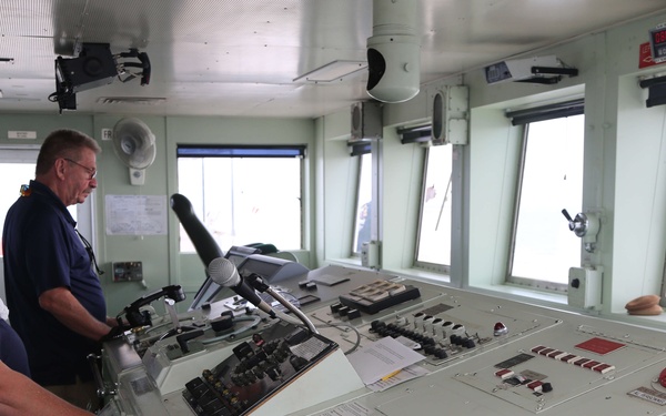 Two individuals stand at the helm of a ship's bridge, facing a large control console with numerous instruments, switches, and navigation equipment. A man in a blue shirt and a woman in a dark top with sunglasses are positioned at the control panel, appearing to observe or operate the vessel. The bridge offers forward-facing windows with a view of open water.