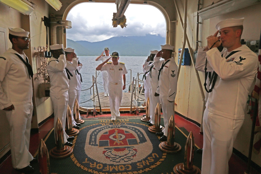 A formal military boarding ceremony is taking place aboard the USNS Comfort, a U.S. Navy hospital ship. Sailors in white dress uniforms are forming a sideboy honor guard, saluting as a senior officer (identifiable by gold shoulder boards) boards the vessel. The scene is framed by a large arched gangway opening with a mountainous coastal landscape visible in the background.