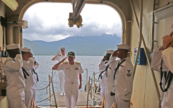A formal military boarding ceremony is taking place aboard the USNS Comfort, a U.S. Navy hospital ship. Sailors in white dress uniforms are forming a sideboy honor guard, saluting as a senior officer (identifiable by gold shoulder boards) boards the vessel. The scene is framed by a large arched gangway opening with a mountainous coastal landscape visible in the background.