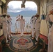 A formal military boarding ceremony is taking place aboard the USNS Comfort, a U.S. Navy hospital ship. Sailors in white dress uniforms are forming a sideboy honor guard, saluting as a senior officer (identifiable by gold shoulder boards) boards the vessel. The scene is framed by a large arched gangway opening with a mountainous coastal landscape visible in the background.