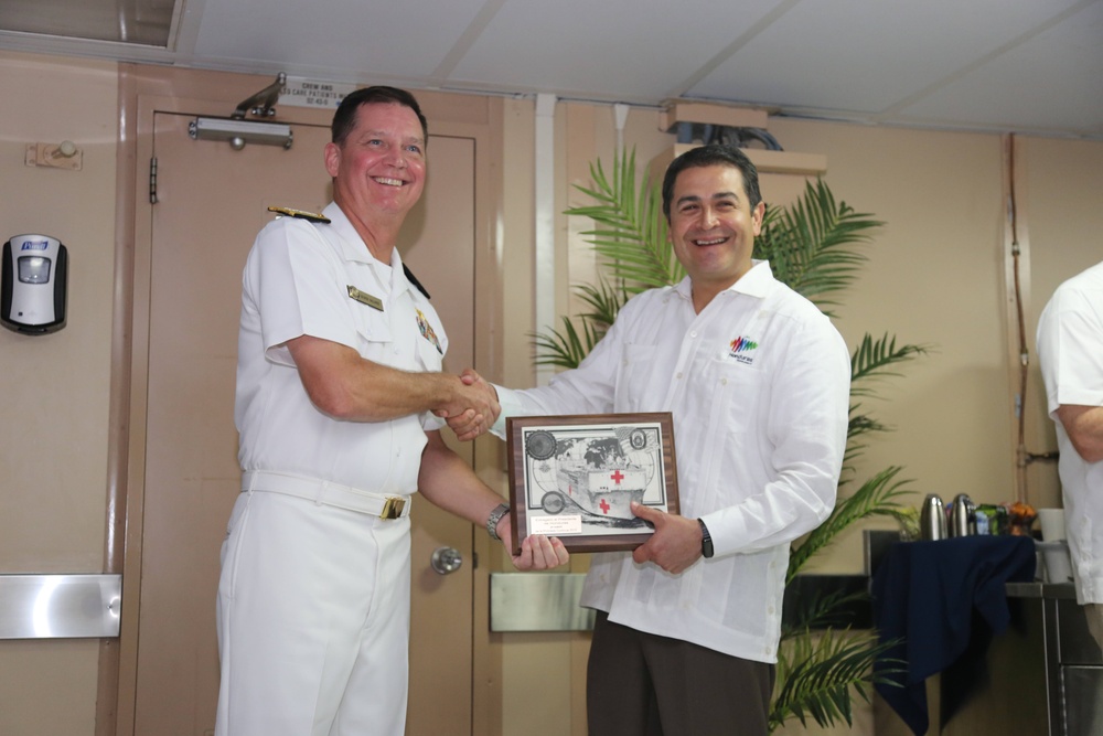 Two men are shaking hands while jointly holding a commemorative plaque in what appears to be an indoor naval or official setting. The man on the left is wearing a white naval dress uniform with gold insignia, while the man on the right is wearing a white guayabera-style shirt with a colorful logo. The plaque features a Red Cross emblem and appears to be an official recognition or ceremonial gift exchange.