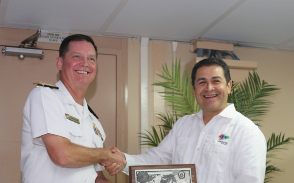 Two men are shaking hands while jointly holding a commemorative plaque in what appears to be an indoor naval or official setting. The man on the left is wearing a white naval dress uniform with gold insignia, while the man on the right is wearing a white guayabera-style shirt with a colorful logo. The plaque features a Red Cross emblem and appears to be an official recognition or ceremonial gift exchange.