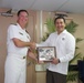 Two men are shaking hands while jointly holding a commemorative plaque in what appears to be an indoor naval or official setting. The man on the left is wearing a white naval dress uniform with gold insignia, while the man on the right is wearing a white guayabera-style shirt with a colorful logo. The plaque features a Red Cross emblem and appears to be an official recognition or ceremonial gift exchange.