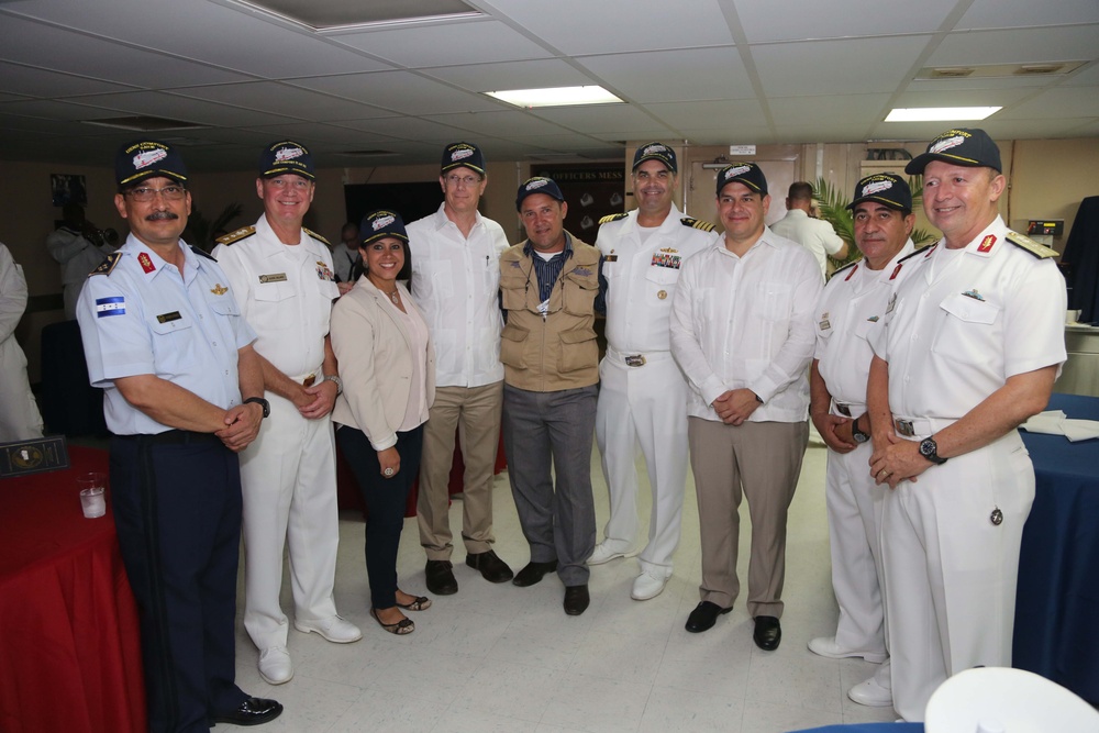 A group of nine individuals pose together for a formal photograph in what appears to be a military mess hall or reception room. Most participants are wearing white naval/military uniforms with matching dark caps, while a few are in civilian attire. The setting suggests an official military gathering or ceremony, likely involving multinational naval personnel.