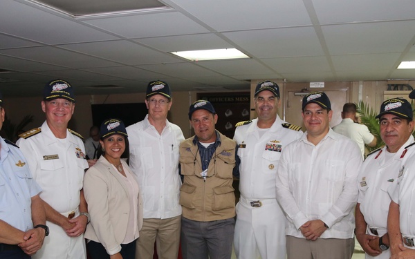 A group of nine individuals pose together for a formal photograph in what appears to be a military mess hall or reception room. Most participants are wearing white naval/military uniforms with matching dark caps, while a few are in civilian attire. The setting suggests an official military gathering or ceremony, likely involving multinational naval personnel.