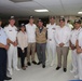 A group of nine individuals pose together for a formal photograph in what appears to be a military mess hall or reception room. Most participants are wearing white naval/military uniforms with matching dark caps, while a few are in civilian attire. The setting suggests an official military gathering or ceremony, likely involving multinational naval personnel.