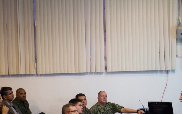 A military briefing or training session is taking place in a conference room setting. A US Marine Corps officer in digital camouflage (MARPAT) uniform is presenting to a mixed audience of military personnel from multiple nations and at least two civilians. Two laptops are visible on a table beside the presenter, suggesting a multimedia presentation.