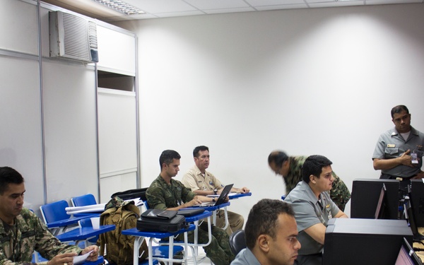 A military training or briefing room with personnel from what appear to be multiple nations or branches, seated at both student desks and computer workstations. Some individuals are in camouflage uniforms while others wear gray or tan military attire, suggesting a joint or multinational exercise environment. The room is equipped with desktop computers on one side and traditional classroom desks on the other.