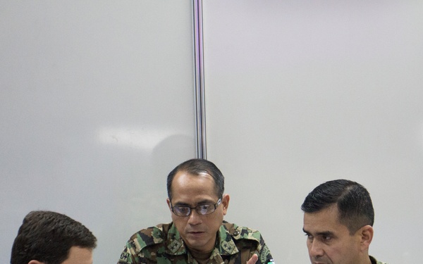 Three military personnel in camouflage uniforms are seated together in what appears to be an indoor briefing or planning session. They are reviewing documents and engaged in active discussion, with the center individual gesturing with his finger as if making a point. The uniforms and patches suggest personnel from multiple nations, indicating a multinational military coordination meeting.