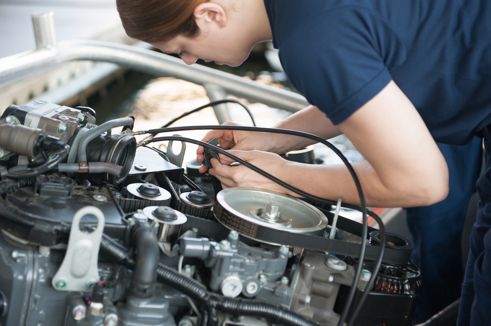 A young mechanic wearing a dark blue shirt is leaning over an open vehicle engine bay, performing hands-on mechanical work. The image focuses on the engine components including belts, pulleys, and various cables. The scene appears to be in an automotive workshop or garage setting.