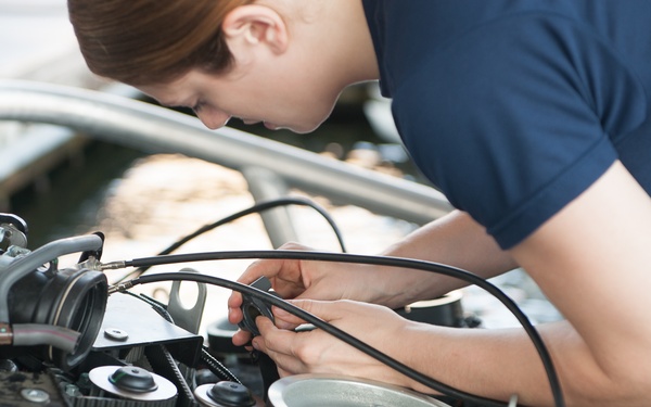 A young mechanic wearing a dark blue shirt is leaning over an open vehicle engine bay, performing hands-on mechanical work. The image focuses on the engine components including belts, pulleys, and various cables. The scene appears to be in an automotive workshop or garage setting.