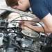 A young mechanic wearing a dark blue shirt is leaning over an open vehicle engine bay, performing hands-on mechanical work. The image focuses on the engine components including belts, pulleys, and various cables. The scene appears to be in an automotive workshop or garage setting.
