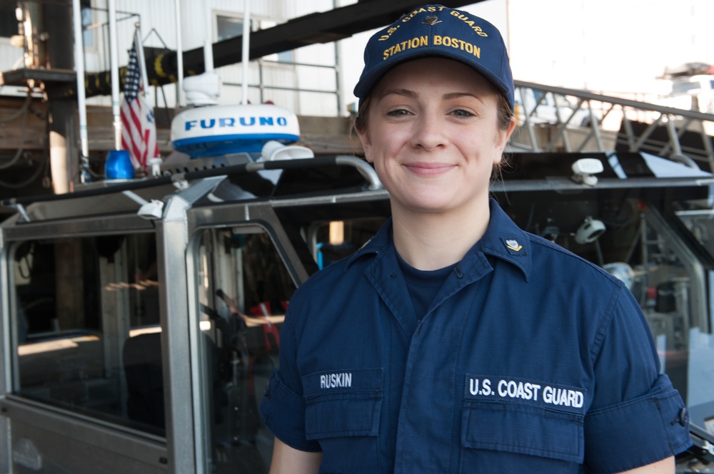 A U.S. Coast Guard member named Ruskin is photographed at Coast Guard Station Boston, smiling at the camera. She is wearing a navy blue Coast Guard uniform with her name tape and branch insignia visible, along with a 'U.S. Coast Guard Station Boston' cap. She is standing in front of a Coast Guard vessel equipped with a Furuno radar dome and an American flag.