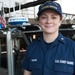 A U.S. Coast Guard member named Ruskin is photographed at Coast Guard Station Boston, smiling at the camera. She is wearing a navy blue Coast Guard uniform with her name tape and branch insignia visible, along with a 'U.S. Coast Guard Station Boston' cap. She is standing in front of a Coast Guard vessel equipped with a Furuno radar dome and an American flag.