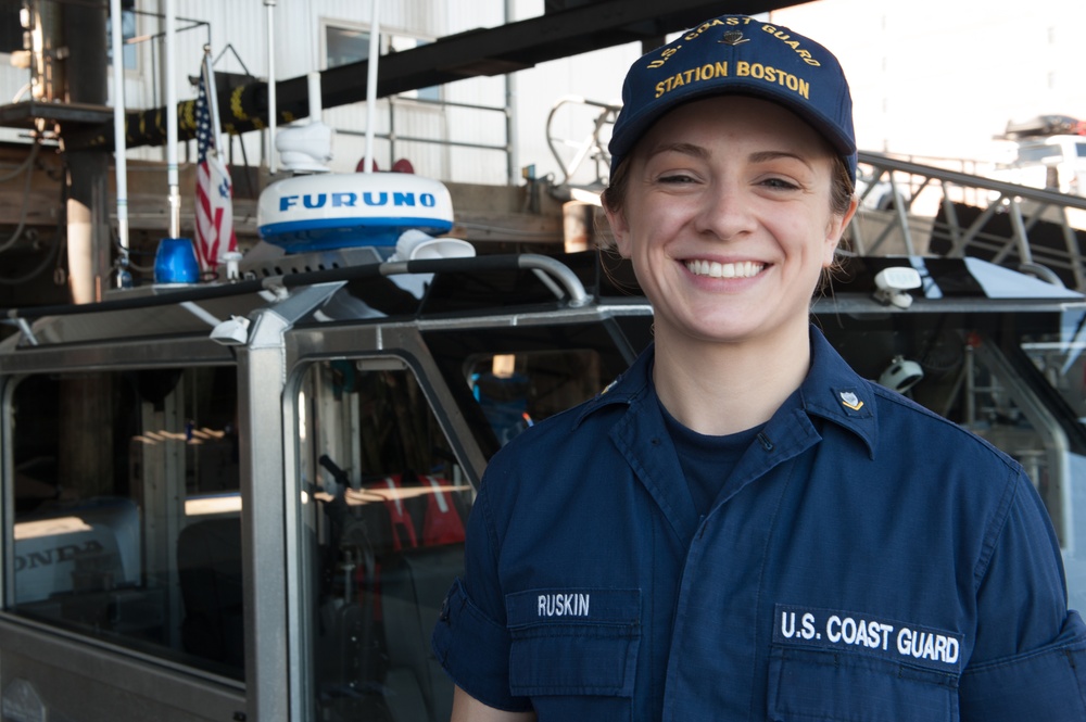 A U.S. Coast Guard member named Ruskin is photographed smiling at Coast Guard Station Boston. She is wearing a navy blue uniform with 'U.S. Coast Guard' and 'RUSKIN' name tape, along with a 'Station Boston' cap. The background shows Coast Guard vessels and equipment, including a Furuno radar dome and an American flag.