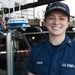 A U.S. Coast Guard member named Ruskin is photographed smiling at Coast Guard Station Boston. She is wearing a navy blue uniform with 'U.S. Coast Guard' and 'RUSKIN' name tape, along with a 'Station Boston' cap. The background shows Coast Guard vessels and equipment, including a Furuno radar dome and an American flag.