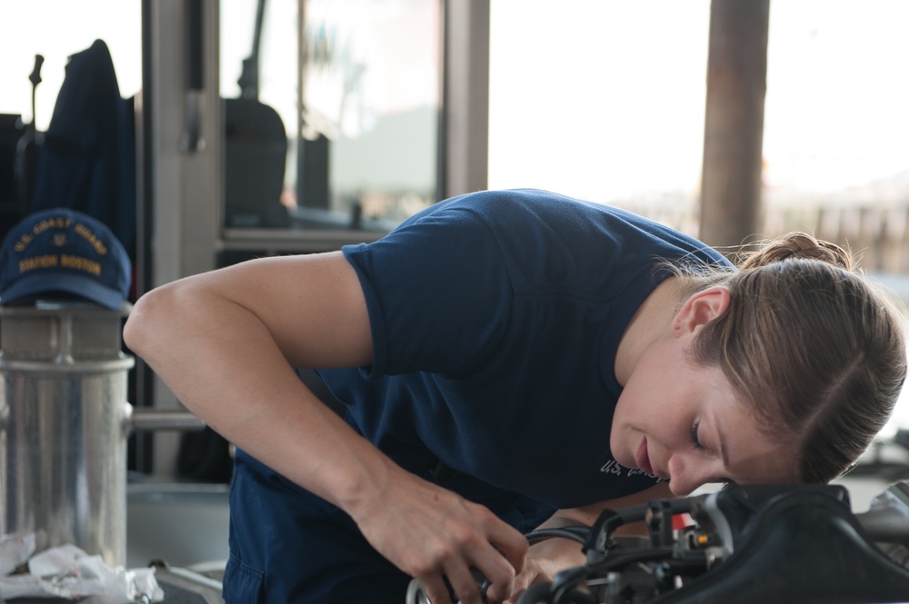 A person wearing a U.S. Coast Guard navy blue t-shirt is leaning over and performing mechanical work on an engine or equipment. In the background, a U.S. Coast Guard 'Station' cap is visible on what appears to be a trash can or container. The setting appears to be aboard a Coast Guard vessel or station facility.