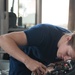 A person wearing a U.S. Coast Guard navy blue t-shirt is leaning over and performing mechanical work on an engine or equipment. In the background, a U.S. Coast Guard 'Station' cap is visible on what appears to be a trash can or container. The setting appears to be aboard a Coast Guard vessel or station facility.